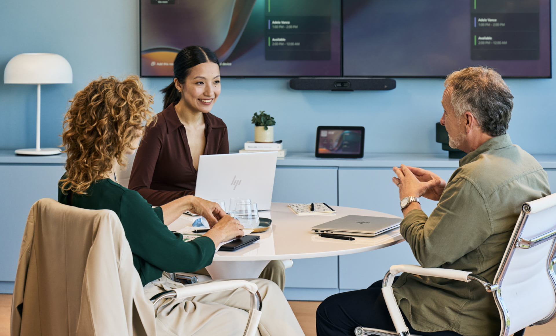 two women and one man in a professional meeting room talking at a round table and using HP laptops in front of an HP Poly large conferencing display
