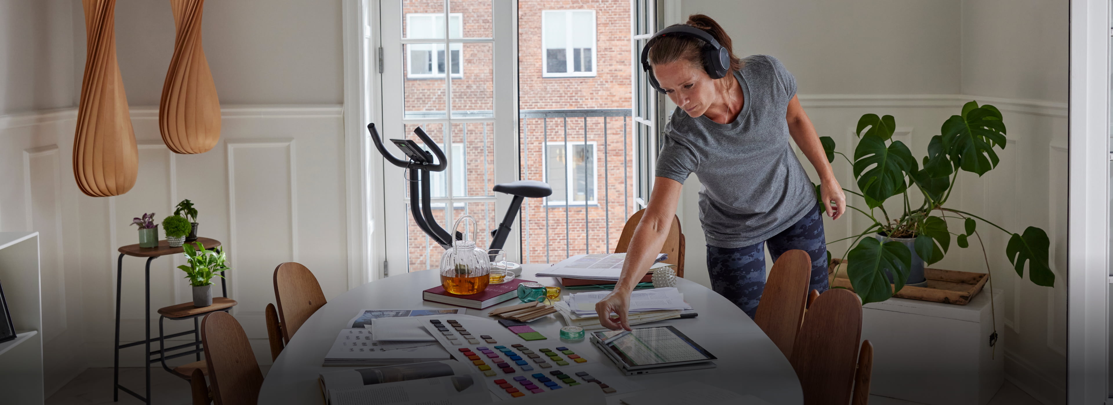 Person using tablet at art-filled table in bright, flexible remote workspace; exercise bike in background.