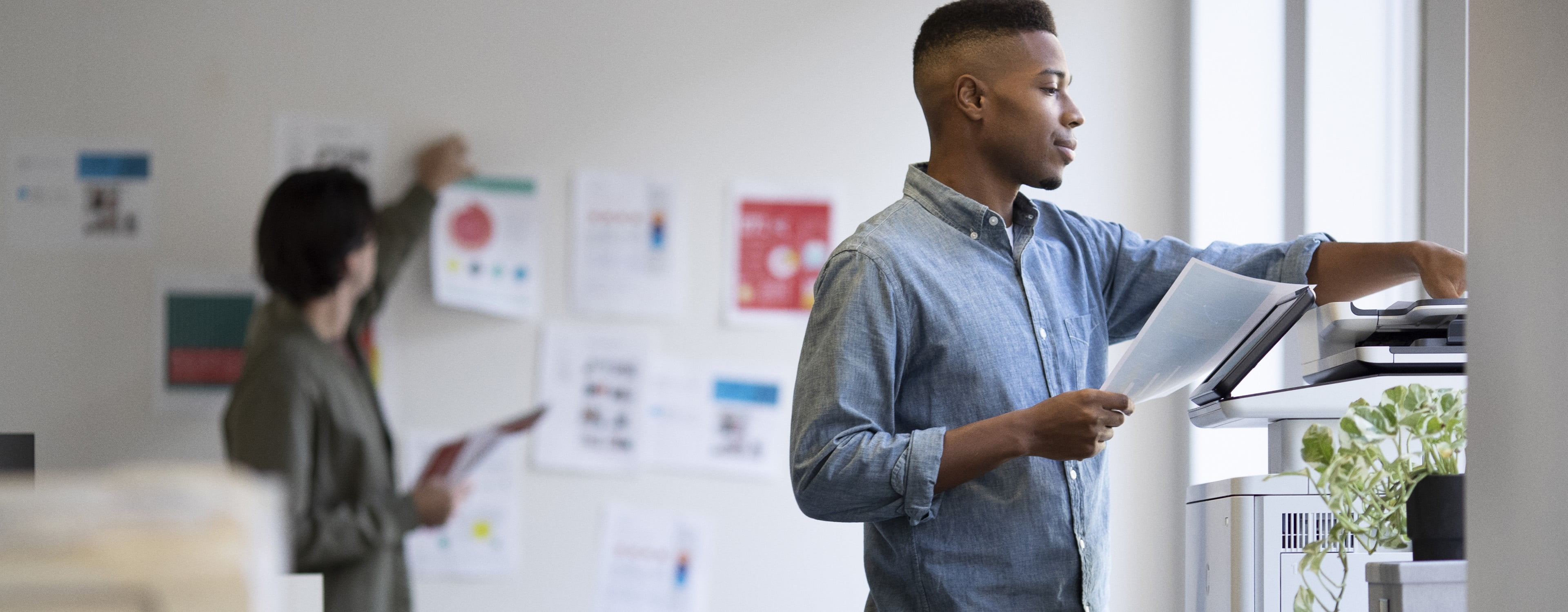 Man in creative office wearing casual attire standing at HP LaserJet Enterprise MFP holding color printed documents with coworker in the rear distance pinning color printe documents to the wall