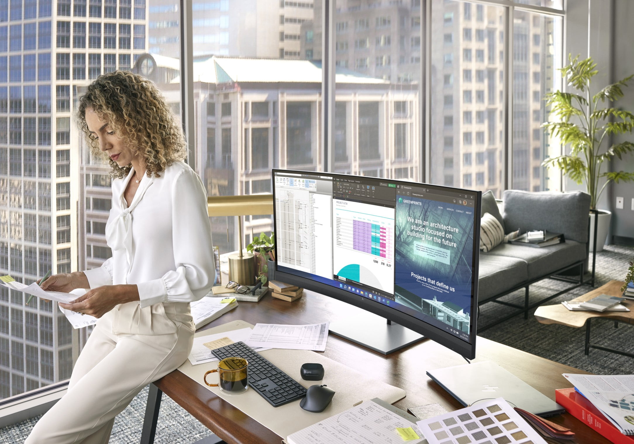 A woman looking at some prints while seated on a desktop aside an HP curved monitor.