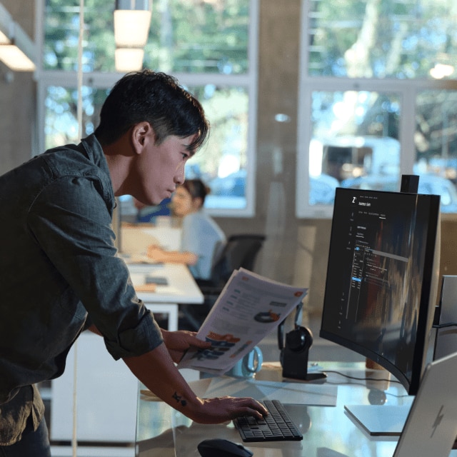 A developer holding reports and tapping on a keyboard in front of a large HP monitor.