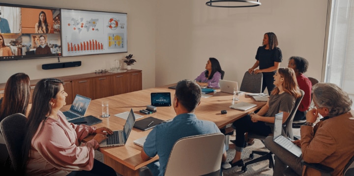  A team meets around a large conference table, with remote colleagues joining from a video feed displayed on an HP wall-mounted monitor. 