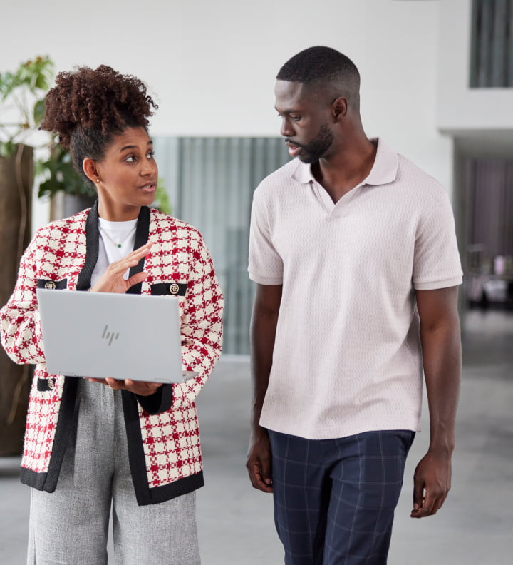 professional man and woman walking side by side, woman holding HP laptop and talking as man listens