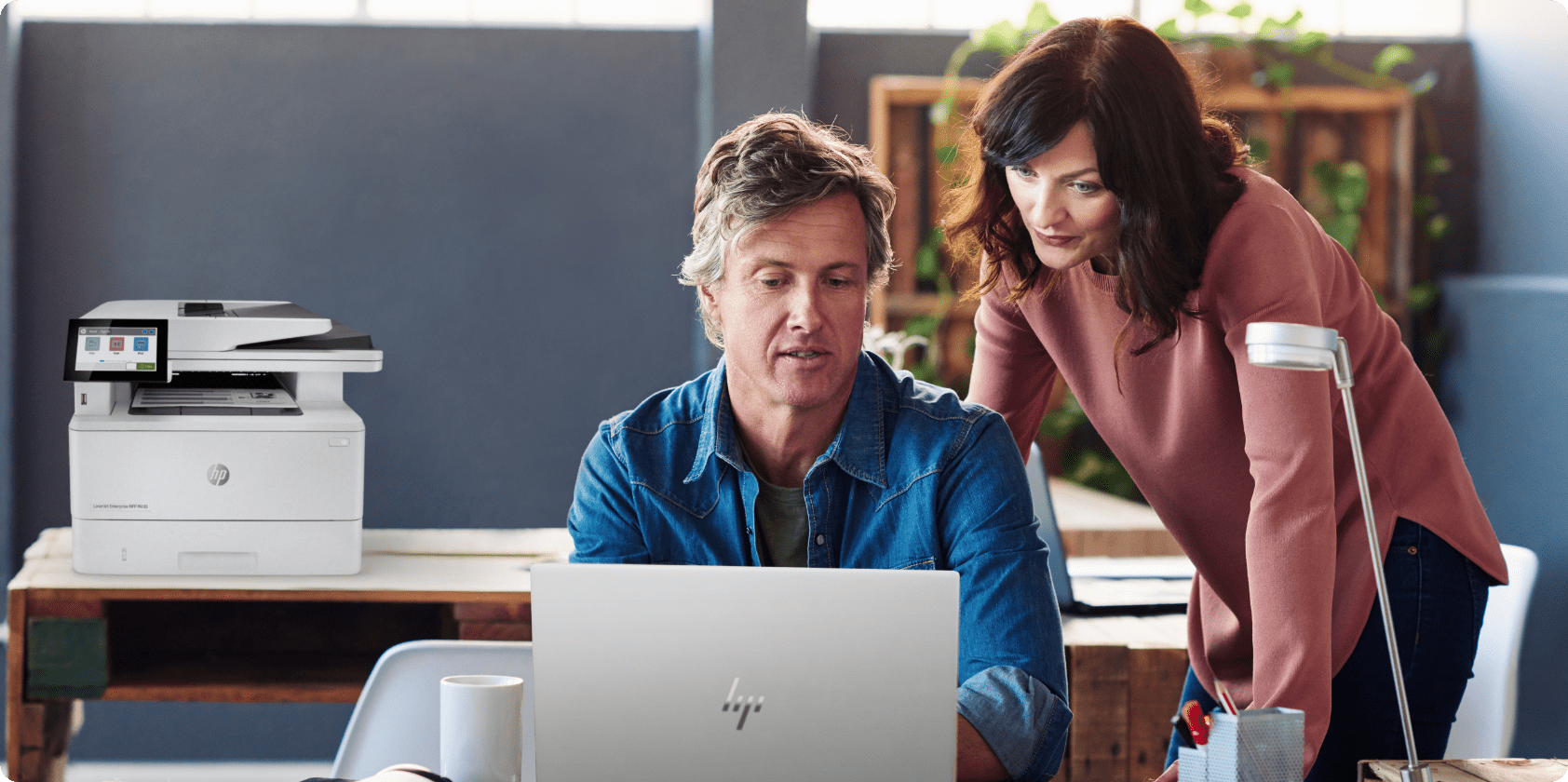 Man seated at desk using HP PC, woman leaning over his shoulder to review the work, HP LaserJet on table behind them