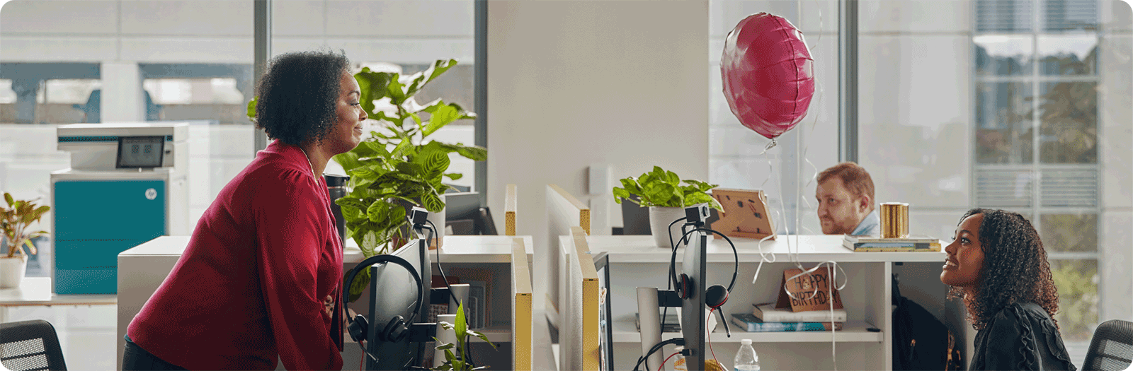 two woman in facing cubicles smiling at one another over their divider wall, with HP monitors and a red balloon